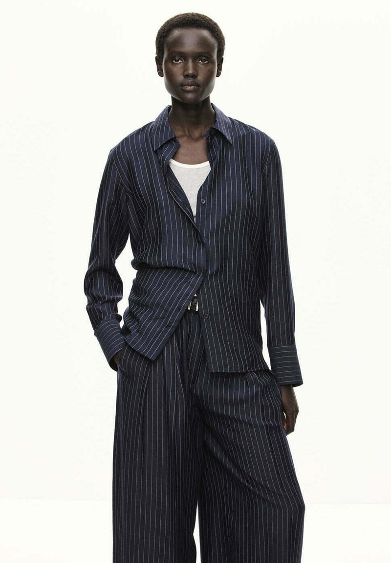 Personne portant une chemise à manches longues rayée foncée et un pantalon large assorti, mains dans les poches, sur un fond clair uni.