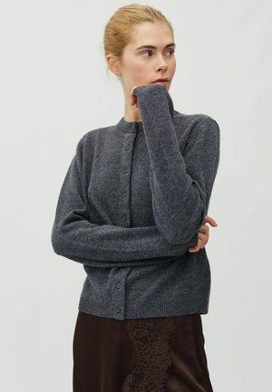 Femme aux cheveux blonds portant un cardigan gris foncé boutonné et une jupe marron avec des détails en dentelle, regardant sur le côté avec une main près du visage.