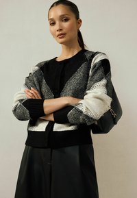 Pletený cardigan v černé a bílé, který má geometrický vzor, kulatý výstřih a knoflíkové zapínání. V kombinaci s černou kabelkou.