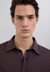 Donkerbruin overhemd met knopen, klassieke kraag, gladde textuur en een enkele knoop bij de halslijn. Eenvoudig ontwerp, zonder patronen.