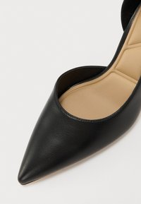 Black high-heeled shoe with a pointed toe and open sides, made of textured leather. The insole is beige, showcasing a smooth surface.
