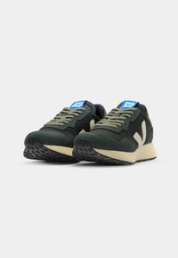 Green and black sneakers with a suede and mesh upper, beige midsole, laces, and blue logo tag. Features textured rubber outsole.