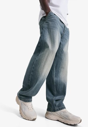 Osoba ubrana w wyblakłe, szerokie, niebieskie dżinsy, białe trampki i białą koszulę, stojąca z rękami w kieszeniach na tle białego tła.