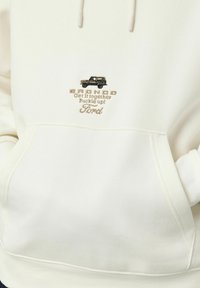 Hoodie blanc avec un graphique Bronco brodé, présentant un texte beige "Remets-toi sur les rails ! Attache ta ceinture !" et "Ford" au-dessus d'un véhicule stylisé noir.