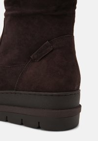 Dunkelbraune Wildlederschuh mit strukturiertem Finish, ausgestattet mit einer dicken schwarzen Gummisohle und einer seitlichen Lasche für einfaches Anziehen.