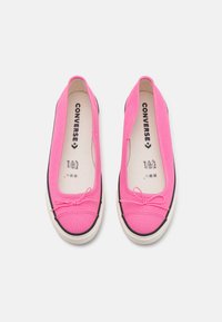 Zapatillas de lona rosas sin cordones con suelas de goma blancas, que presentan detalles en negro y un lazo decorativo en la parte delantera.