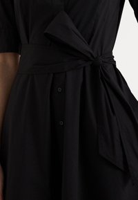 Närbild av en svart klänning med korta ärmar, knäppning framtill och ett brett bälte knutet i en slinga vid midjan.