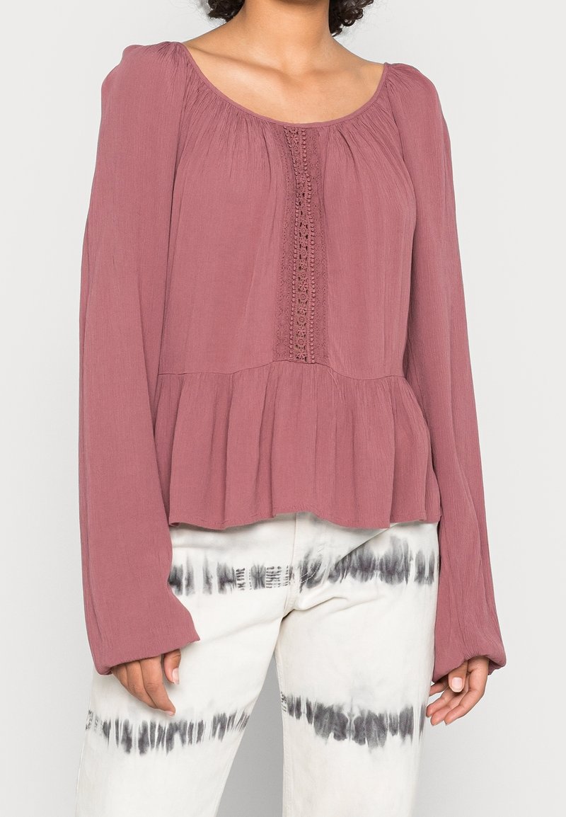 Blouse mauve à manches longues avec détail en dentelle sur le devant, associée à un pantalon tie-dye blanc avec des rayures horizontales noires.
