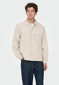 Veste beige boutonnée en tissu léger, dotée d'un col pointu, d'une poche avant et d'une coupe décontractée, associée à un jean bleu foncé.