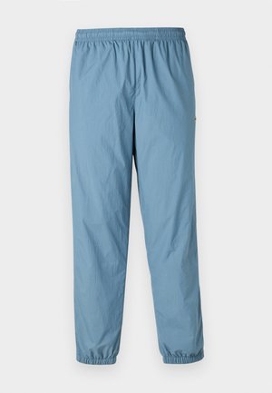 Pantaloni blu, leggeri, con vita elasticizzata, gamba a sigaretta e polsini elasticizzati; presentano un piccolo logo sul lato. Materiale liscio e resistente all'acqua.