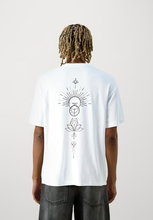 T-shirt en coton blanc avec un grand design graphique noir au dos, représentant un soleil avec des rayons, un lotus et des éléments décoratifs.