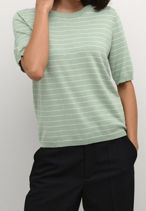 Person trägt einen hellgrünen kurzärmeligen Pullover mit weißen horizontalen Streifen und schwarze Hosen, eine Hand berührt den Hals, die andere steckt in der Tasche.