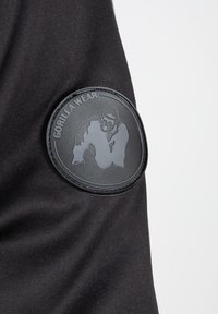Zwarte stof met een rond zwart rubberen embleem met de opdruk "GORILLA WEAR" en een gorilladesign aan de zijkant.