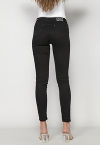 Black skinny jeans with a smooth texture, mid-rise waist, and two back pockets. Worn with transparent high-heeled sandals.