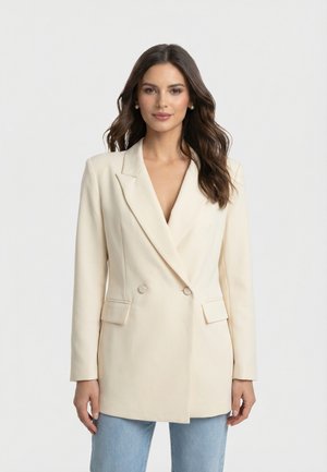 Femme aux longs cheveux noirs portant un blazer crème à double boutonnage sur un jean bleu clair, debout devant un fond gris neutre.