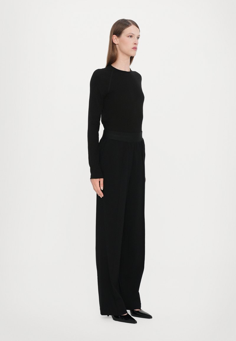 Black long-sleeve top paired with wide-leg black trousers. Smooth fabric, minimal design, and a fitted waistband with sleek black shoes.