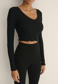 Schwarzes, geripptes Langarm-Crop-Top mit V-Ausschnitt und Volantsaum. Der Stoff ist weich und hat eine figurbetonte Silhouette, die die Taille betont.