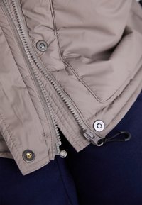 Light beige padded jacket with a double zipper, silver hardware, and drawstring. Features quilted texture and contrasting navy fabric below.