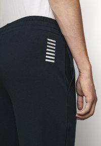 Pantalons de jogging bleu marine avec poches latérales, taille élastique et détails rayés argentés à l'arrière. Texture de tissu douce et lisse.