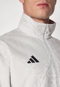 Giacca bianca con zip, caratterizzata da un motivo floreale testurizzato e un colletto alto. Presenta un logo Adidas nero sul petto.