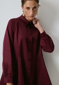 Woman with short curly hair wearing gold drop earrings and a loose-fitting burgundy button-up dress, posing against a plain light background.