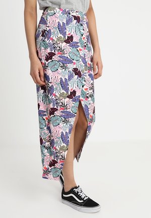 Osoba ubrana w pastelową kwiecistą spódnicę z przednim rozcięciem i czarno-białe sneakersy na sznurowadła, stojąca na tle jednolitego tła.