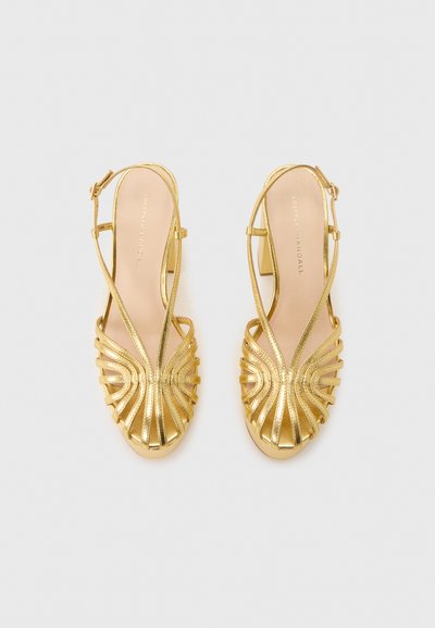 Ζευγάρι χρυσά μεταλλικά πέδιλα ανοιχτού δακτύλου με λουράκι στο πίσω μέρος και καμπυλωτό σχέδιο λουριού, με χοντρά τακούνια σε ανοιχτό γκρι φόντο.