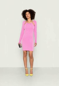 Vestido ajustado de mangas longas em rosa brilhante, apresentando detalhes com ombro à mostra. Usado com sapatos de salto amarelos e segurando uma mala preta.