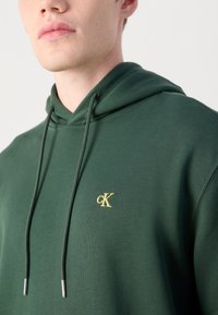 Zelená mikina s kapucňou vyrobená z bavlny, s predným klokaním vreckom, kapucňou so šnúrkami a malým žltým logom "CK" na hrudi.