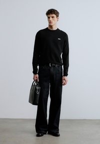 Musta pitkähihainen paita, jossa pieni logo, yhdistetty tummiin leveälahkeisiin farkkuihin, mustaan vyöhön ja harmaaseen nahkalaukkuun. Sileät tekstuurit, minimalistinen muotoilu.