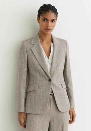 Blazer marrone con trama, bavero a lancia, chiusura con un bottone, due tasche frontali e taglio su misura, indossato sopra una camicetta bianca.