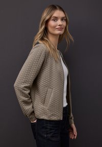 Beige gesteppte Jacke mit Reißverschluss und zwei Seitentaschen. Der Stoff hat ein strukturiertes Rautenmuster, kombiniert mit einem weißen Hemd und dunklen Jeans.
