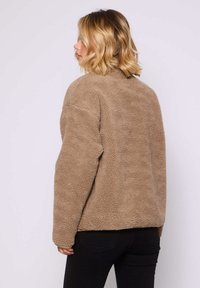 Kobieta z blond włosami, ubrana w jasnobrązową, teksturowaną kurtkę z polaru i czarne spodnie, odwrócona, by pokazać tył kurtki.