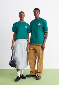 Teal bomulds T-shirts med logo, en lys grå plisseret nederdel, beige kordfløjlsbukser og sorte sko på et grønt gulv.