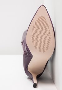 Zapato de tacón alto morado con punta afilada y parte superior lisa. La suela es beige con un patrón de agarre texturizado y un tacón delgado.