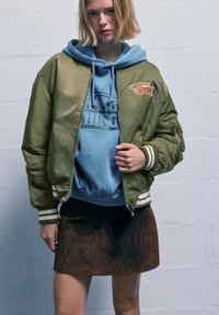 Bomber verde oliva con polsini a righe bianche, indossato sopra una felpa con cappuccio azzurro chiaro. Il modello lo abbina a una gonna in camoscio marrone.