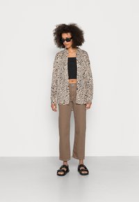 Camisa de botão às riscas zebra bege e preta, top preto curto, calças retas castanhas e sandálias pretas numa modelo com cabelo encaracolado.