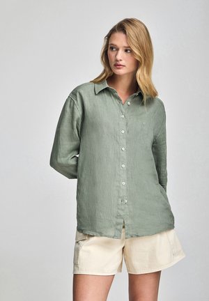 Camicia di lino verde chiaro con bottoni frontali, colletto e maniche lunghe, abbinata a pantaloni corti beige. Tessuto testurizzato, design casual.