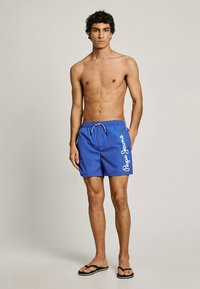 Μπλε μαγιό βερμούδα από ελαφρύ υλικό, με κορδόνι στη μέση και λευκό λογότυπο στο πλάι. Φορεμένα με μαύρες σαγιονάρες.