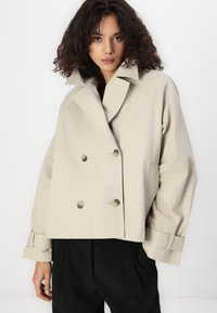 Beige double-breasted jacket with a wide collar, large gold buttons, and rolled cuffs, paired with black trousers against a white background.