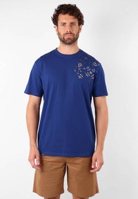 T-shirt en coton bleu avec des manches courtes, un col rond et un détail imprimé floral doré sur l'épaule gauche. Associé à un short marron.