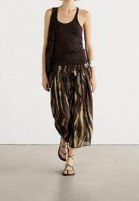 Vrouw in een zwarte tanktop, met een losse broek met bruine en gouden strepen, zwarte sandalen, die een zwarte schoudertas draagt en een zilveren armband om heeft.