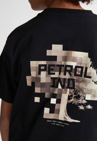 Zwarte katoenen T-shirt met een grafische print met de tekst "PETROL IND" en een pixelontwerp van een boom in beige en bruintinten.