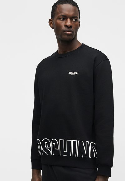 Άνδρας που φοράει μαύρο φούτερ με το λογότυπο "MOSCHINO ACTIVE" στο στήθος και μεγάλο κείμενο "MOSCHINO" εν μέρει ορατό κοντά στη μέση.