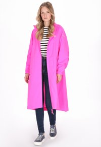 Hosszú, élénk rózsaszín esőkabát kapucnival, rugalmas mandzsettákkal és elülső zsebekkel, fekete-fehér csíkos ing és sötét farmer felett viselve.
