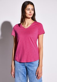 Růžové tričko s krátkým rukávem a véčkovým výstřihem vyrobené z lehké tkaniny s volným střihem a hladkou strukturou, v kombinaci se světle modrými džíny.