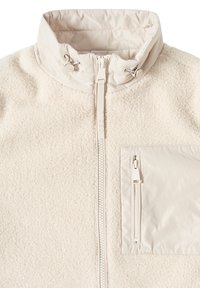 Veste en polaire beige clair avec un col haut, fermeture éclair sur le devant et une poche poitrine en tissu lisse contrastant avec zip. Tissu texturé sur l'ensemble.