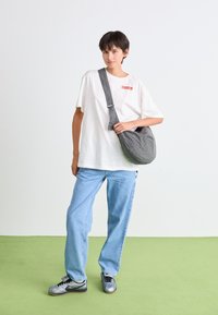 Gray plaid sling bag made of soft fabric, curved shape, featuring a wide strap. Model wearing a white t-shirt and light blue jeans.