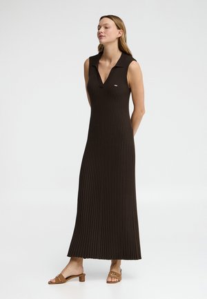 Kobieta stoi z rękami za plecami, ubrana w długą, czarną, prążkowaną sukienkę bez rękawów i brązowe sandały na obcasie, na jednolitym tle.