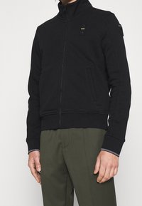 Svart zip-up sweatshirt med hög krage, tillverkad av mjukt material. Har sidofickor och en distinkt patch på bröstet.